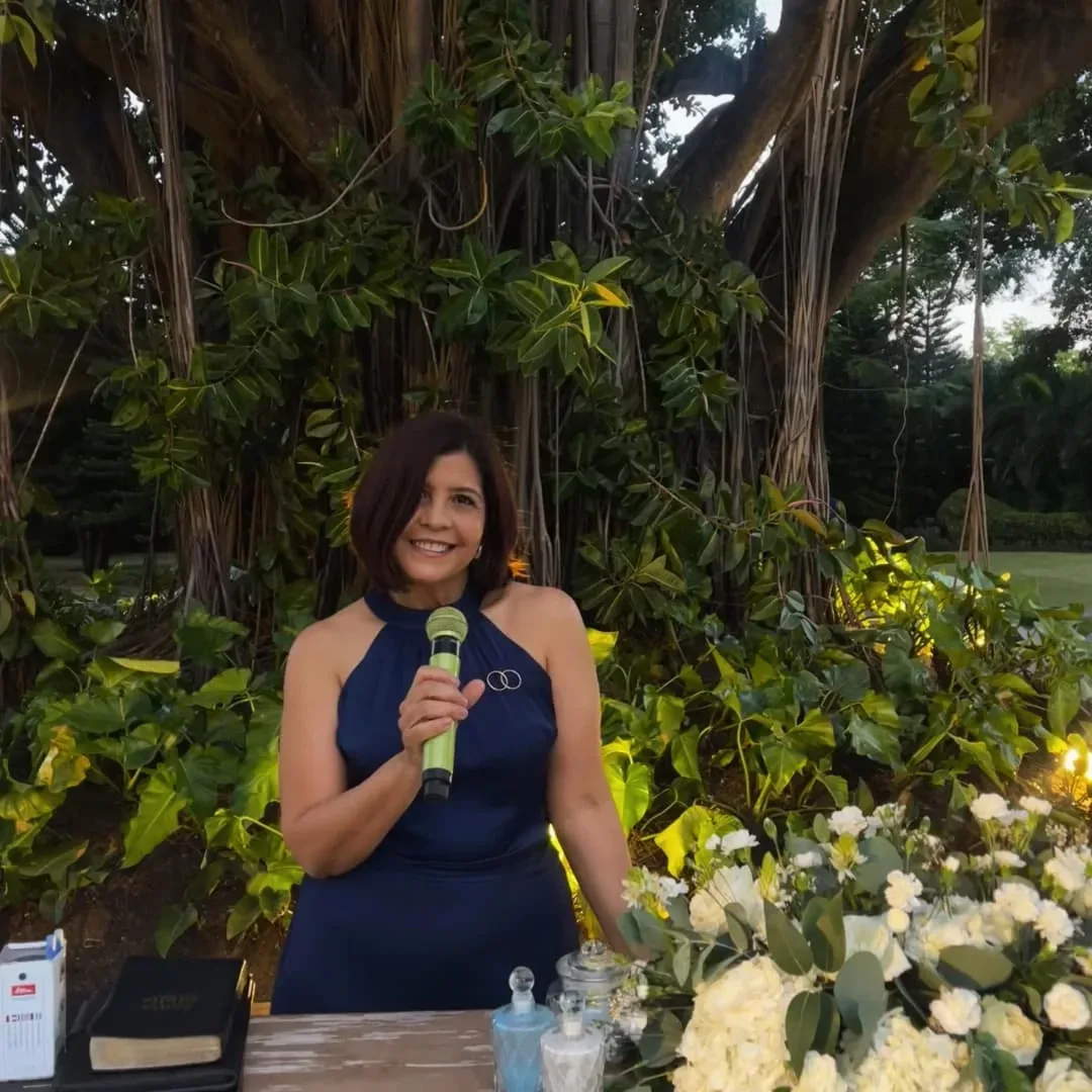 Luz Polanco, de vestido azul y micrófono en mano, frente a un árbol frondoso y flores blancas; visitó Tarahumara