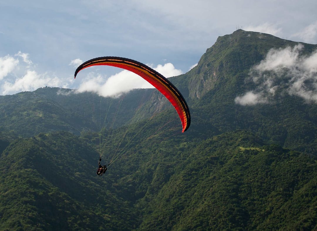 Parapente rojo y negro sobrevolando las montañas verdes de El Ávila entre nubes, cerca de Galipán.