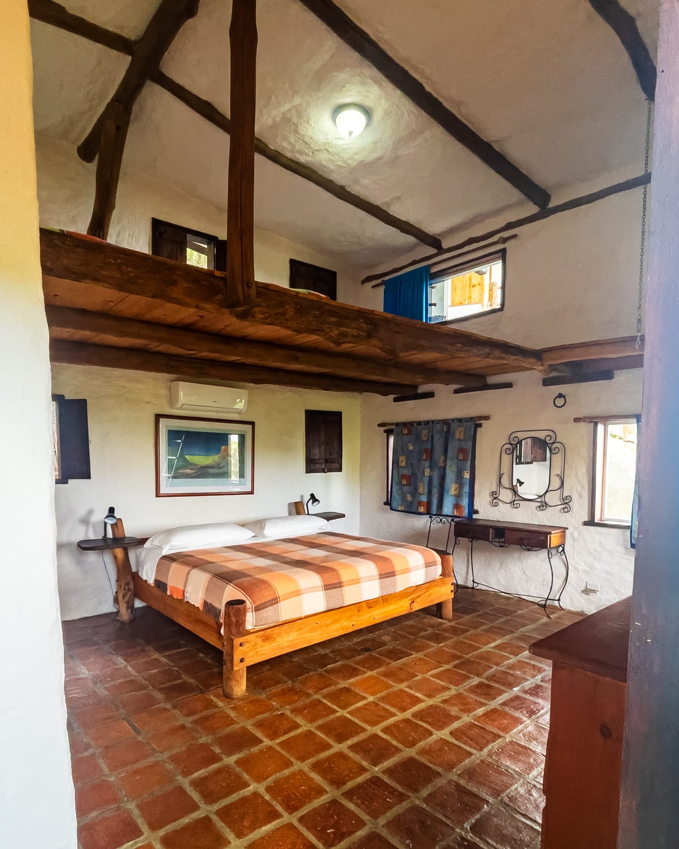Dormitorio de doble altura con vigas, cama king de cobija a cuadros, peinadora y piso de terracota
