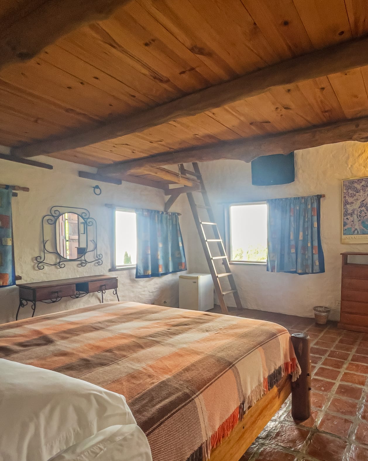 Dormitorio de techo de tablones con cama king de manta a rayas, tocador de hierro y la escalera al altillo