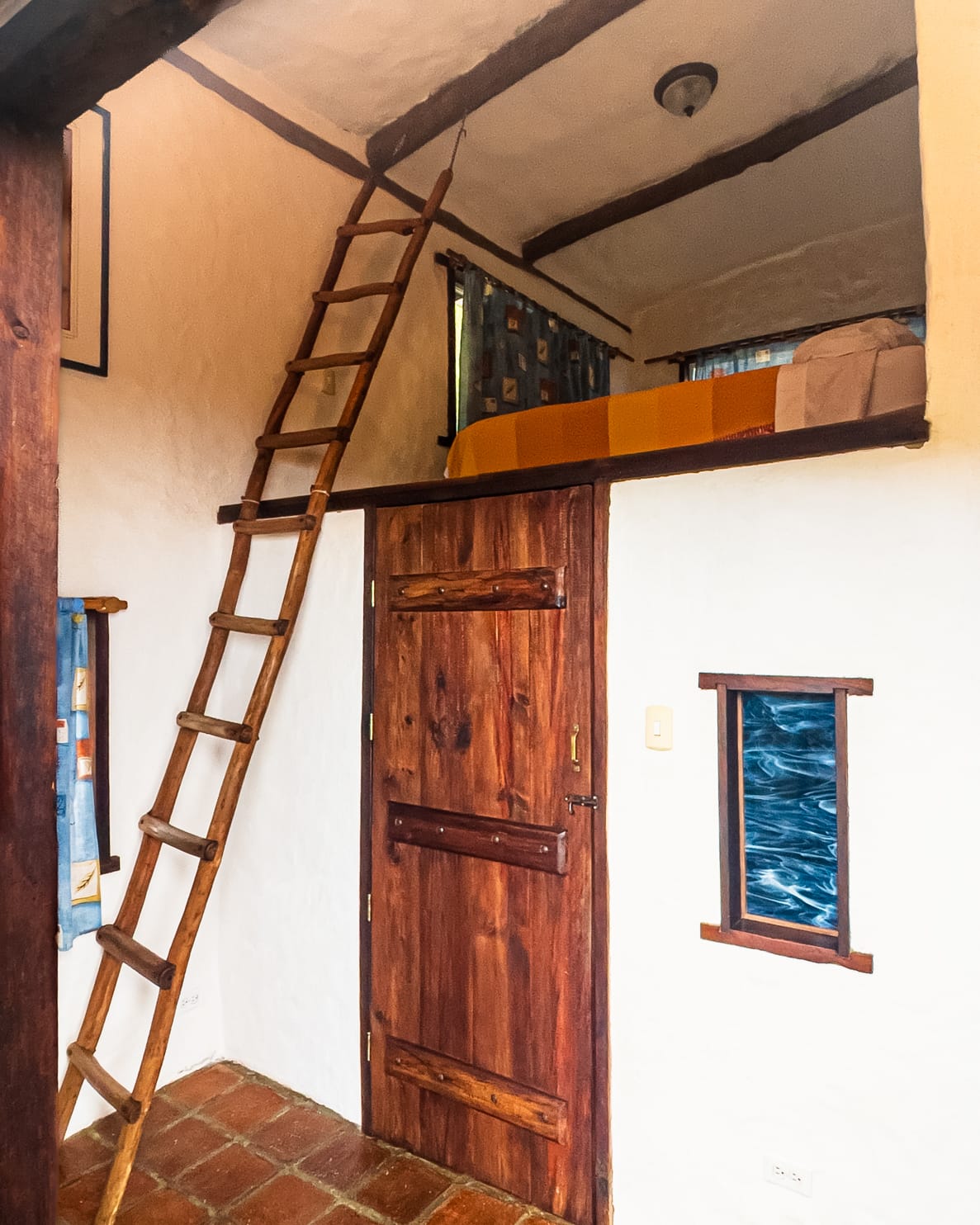 Escalera de troncos que sube al altillo con una cama de cobija anaranjada, sobre la puerta rústica de madera