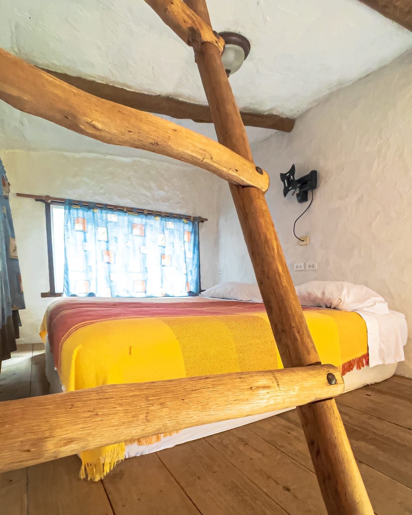 Entre los travesaños de la escalera asoma una cama con cobija amarilla y roja en el piso de madera del altillo