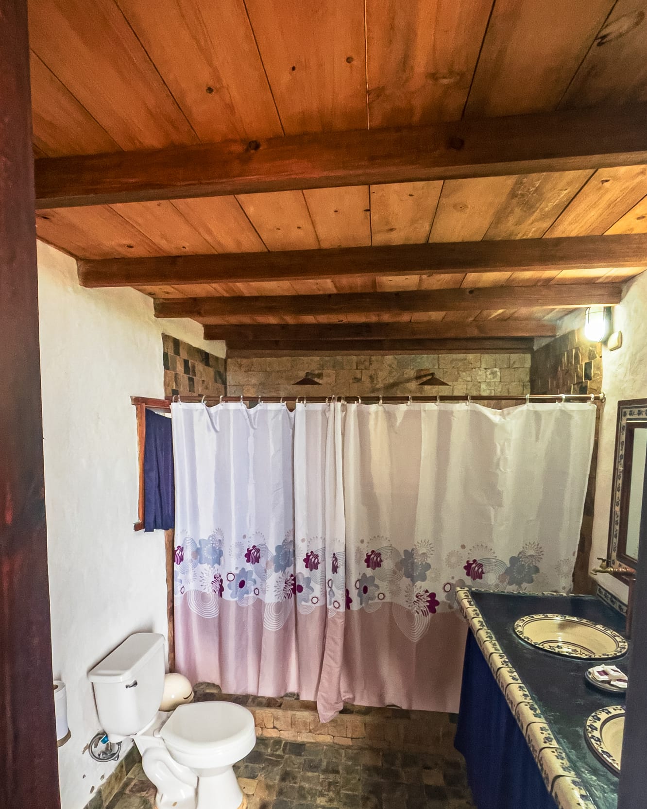 Baño con techo de madera, cortinas floreadas en la ducha doble y dos lavamanos de cerámica pintada