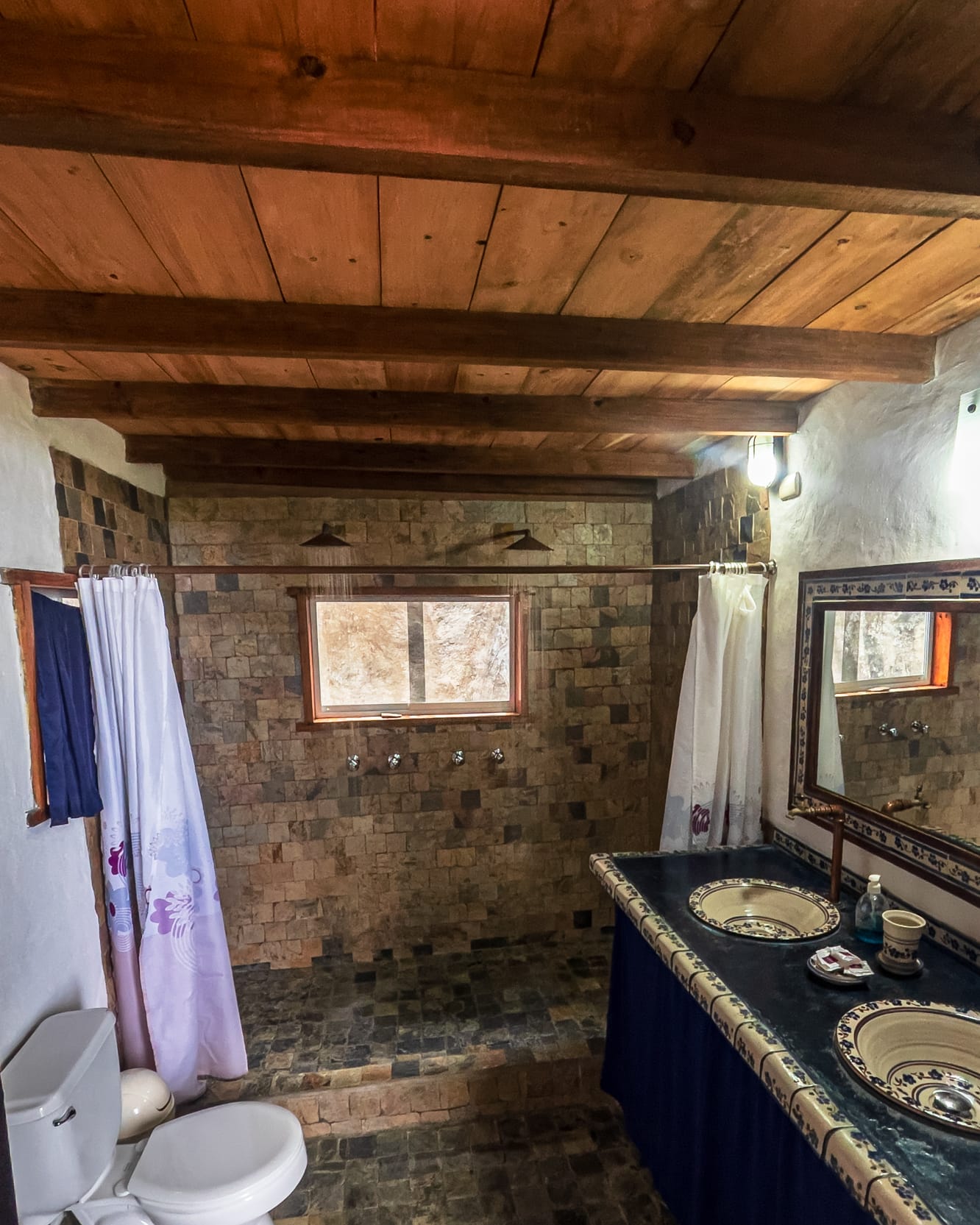 La ducha doble de pared de piedra con su ventanita, frente al mesón azul de dos lavamanos artesanales
