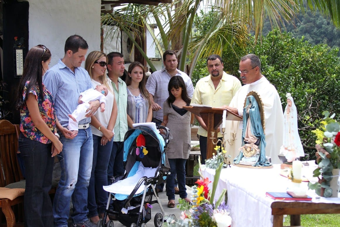 Familia con el bebé en brazos junto al altar con la Virgen mientras el sacerdote lee, en el jardín de Posada Tarahumara.