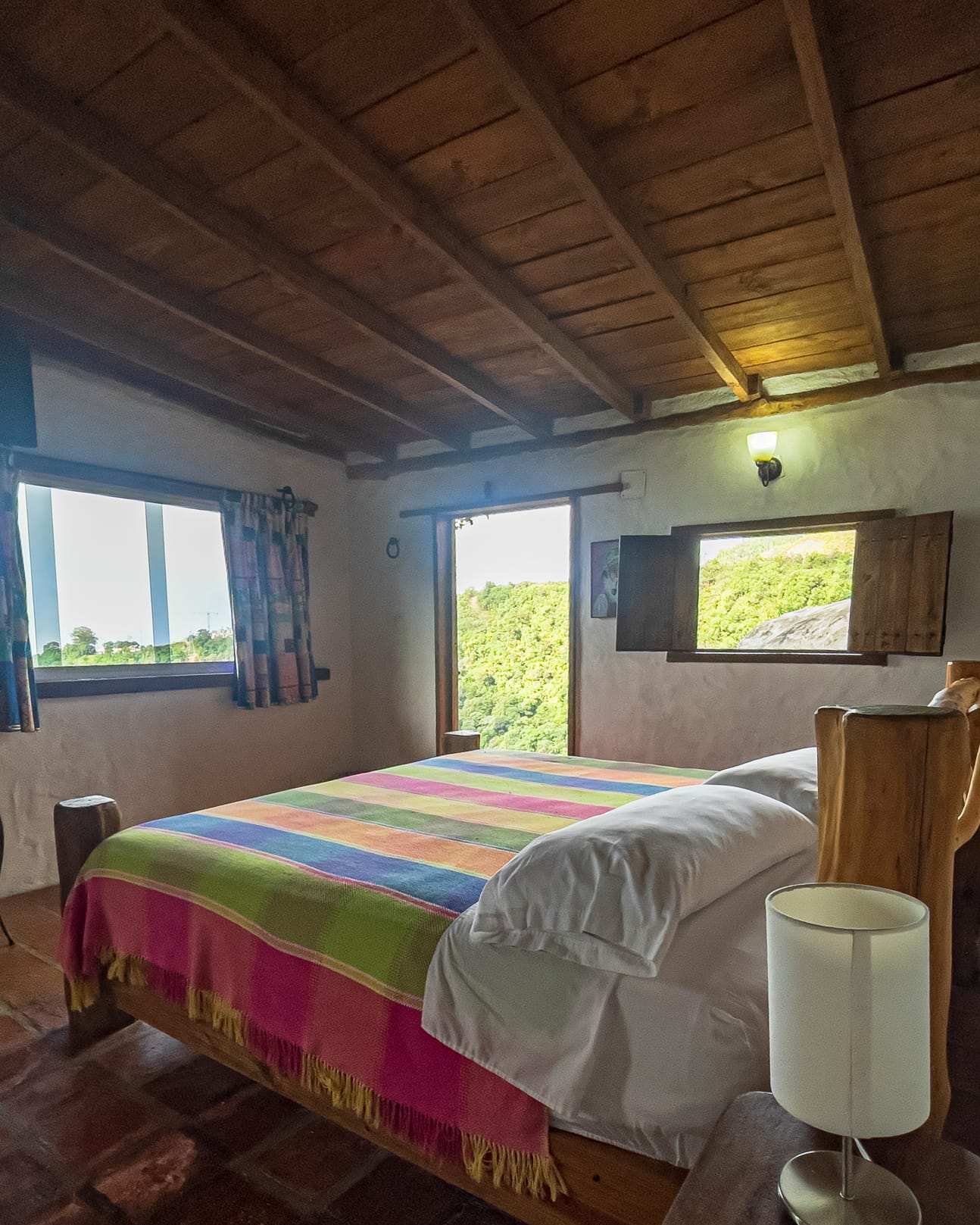 Cama king con cobija de rayas multicolor bajo el techo de vigas, y dos ventanas abiertas al verde de la montaña