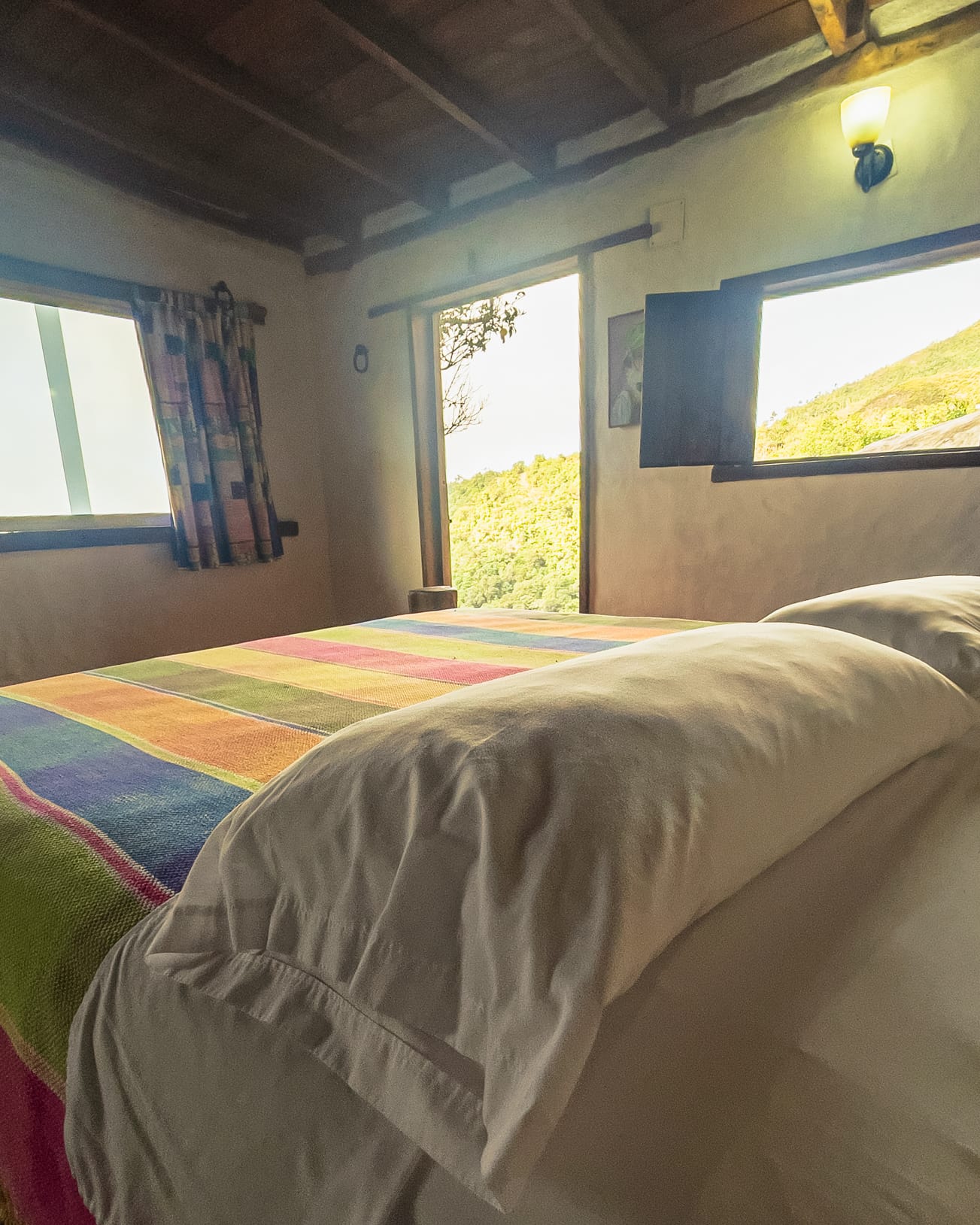 Almohadas blancas sobre el tejido de colores, con la ventana de postigos abierta hacia la ladera boscosa