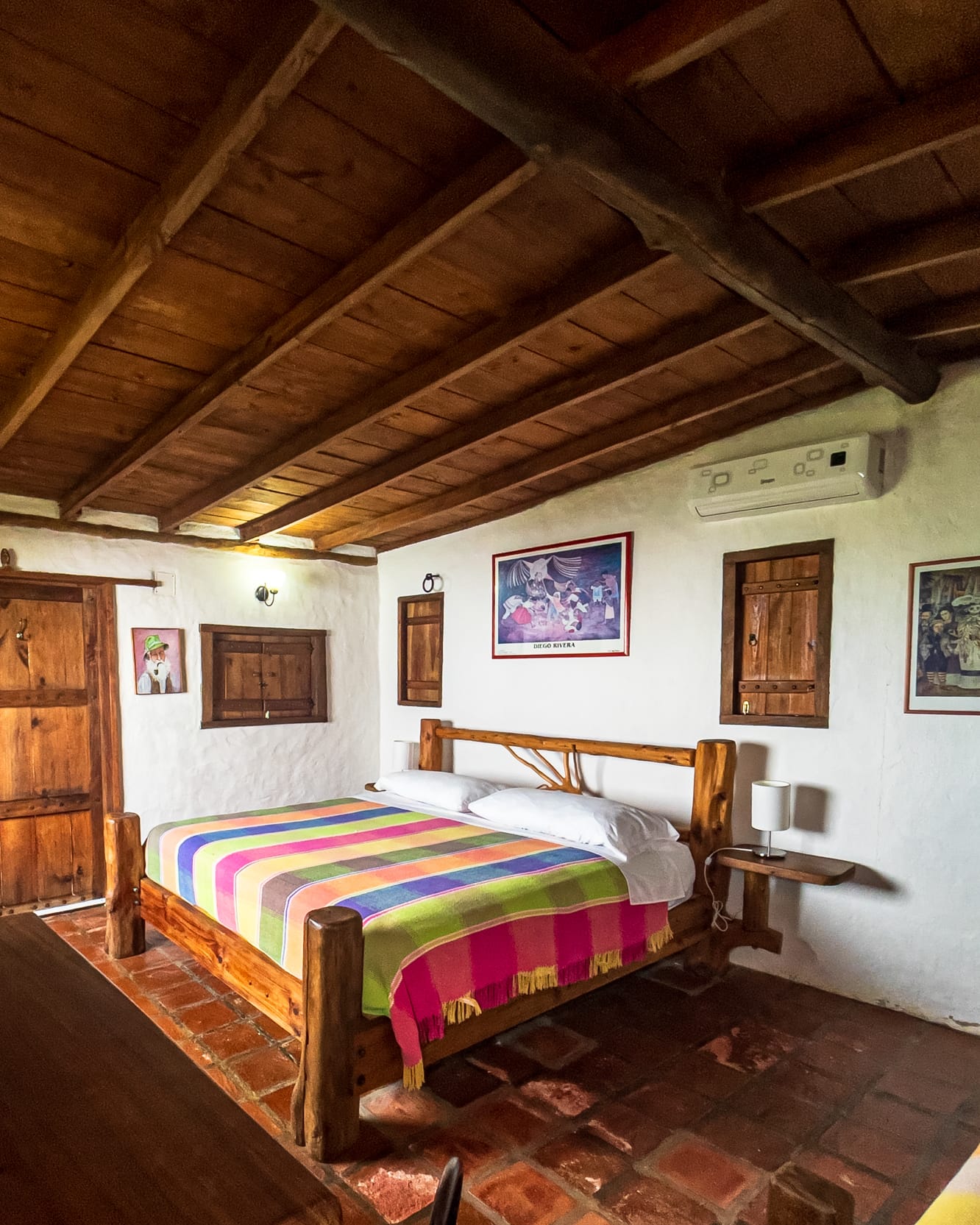 Dormitorio de paredes encaladas y piso de terracota, con cama de troncos, cobija de rayas y aire acondicionado