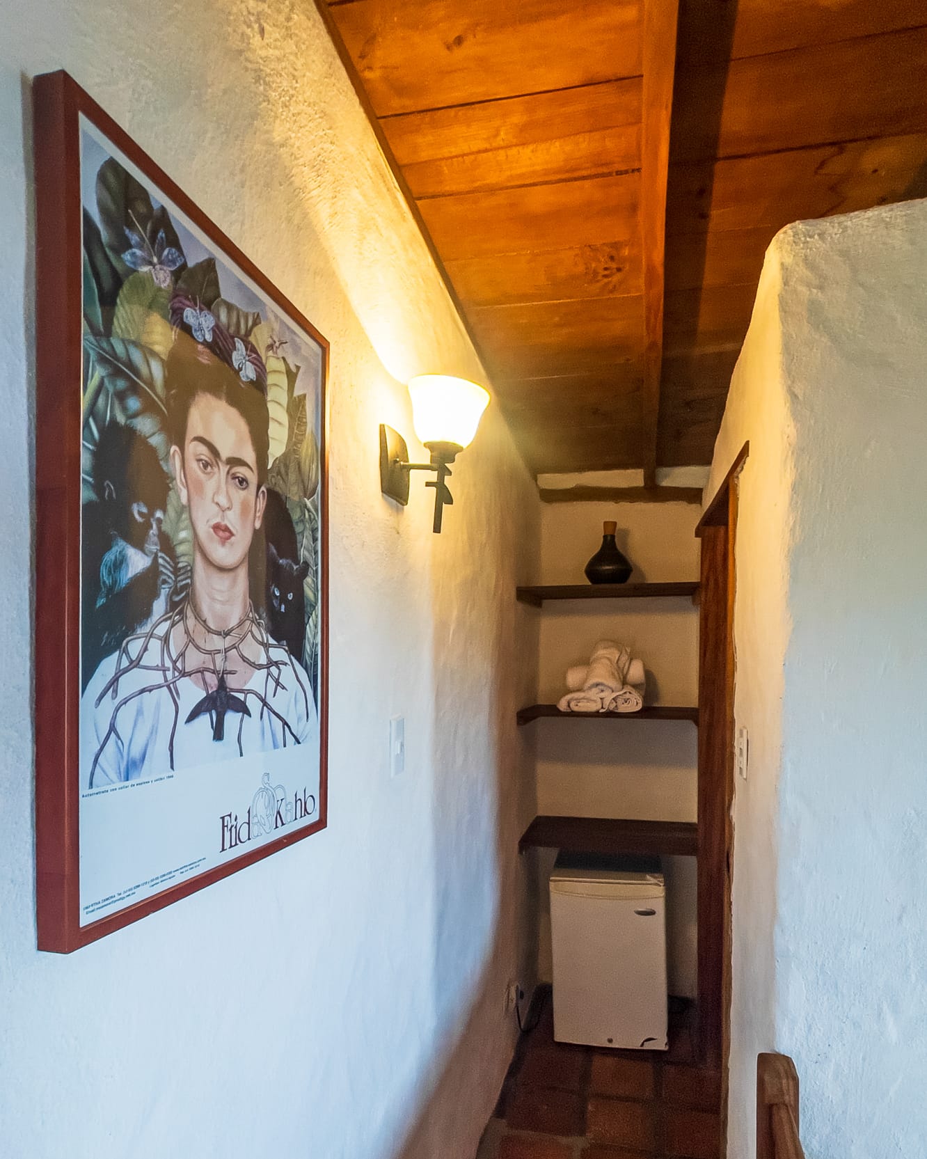 Pasillito con un afiche de Frida Kahlo, aplique encendido y repisas con toallas sobre la nevera ejecutiva
