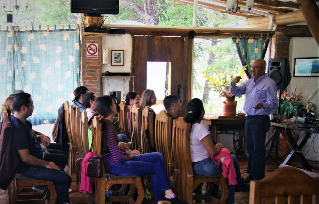 Ponente de pie frente a un grupo sentado en sillas de madera, en el salón rústico de Posada Tarahumara, Galipán.