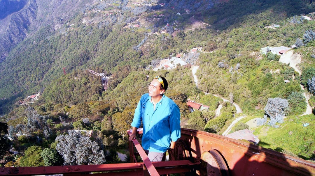 Hombre de camisa azul sobre la vieja torre de hierro del teleférico, con las casas y montañas de Galipán al fondo