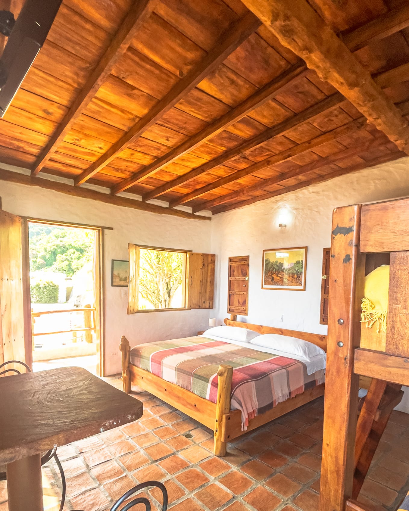 Techo de vigas a la vista sobre la cama matrimonial con cubrecama de cuadros y piso de terracota