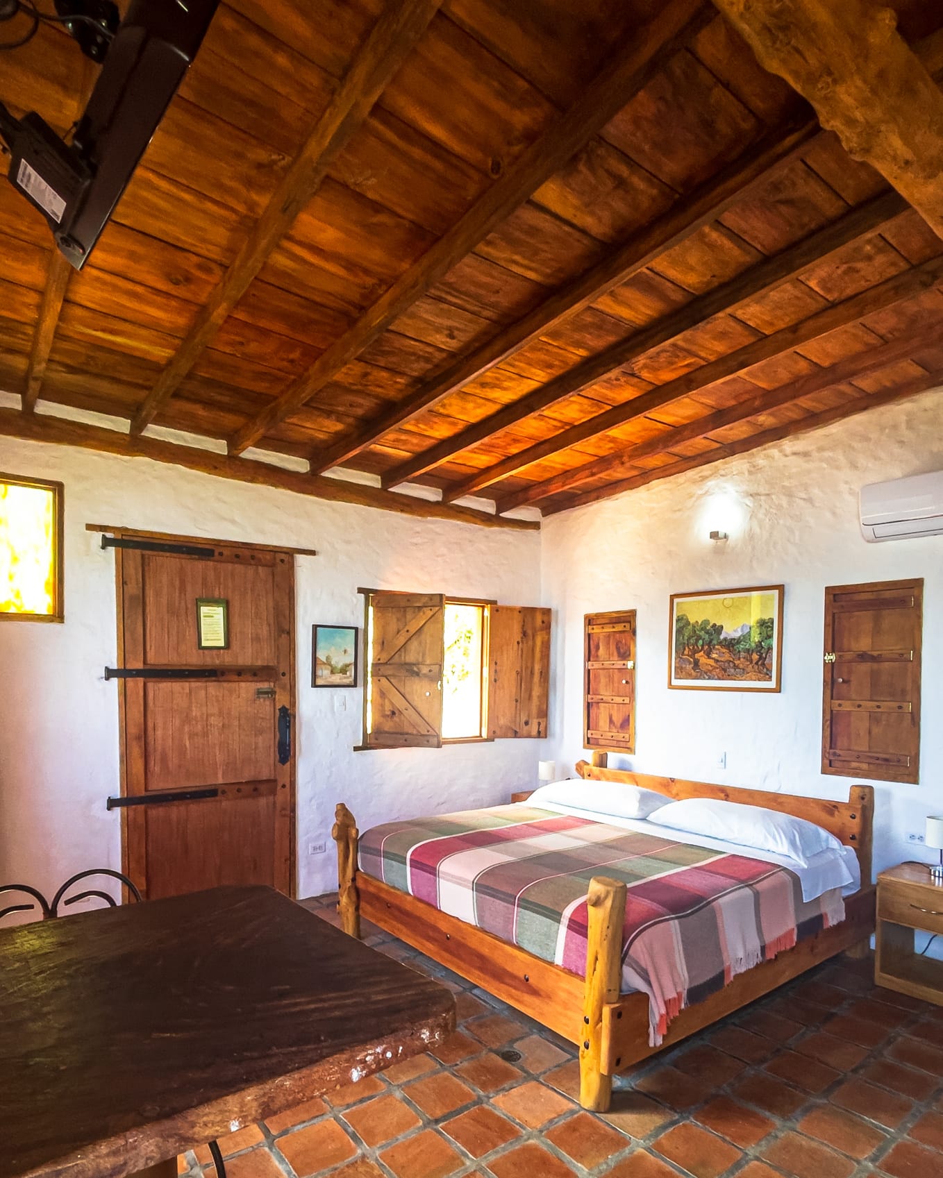 Habitación de paredes encaladas con puerta rústica de herrajes negros, aire acondicionado y cama de madera