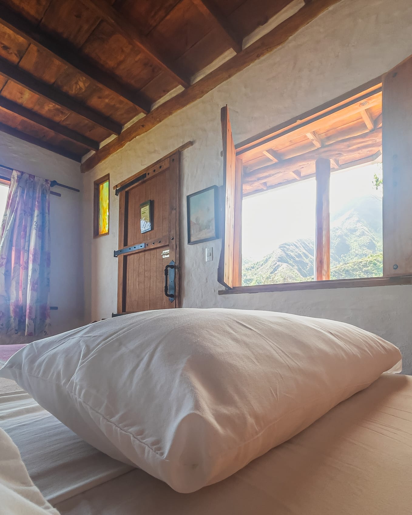 Almohada blanca en primer plano, con la puerta rústica al fondo y el cerro asomado en la ventana