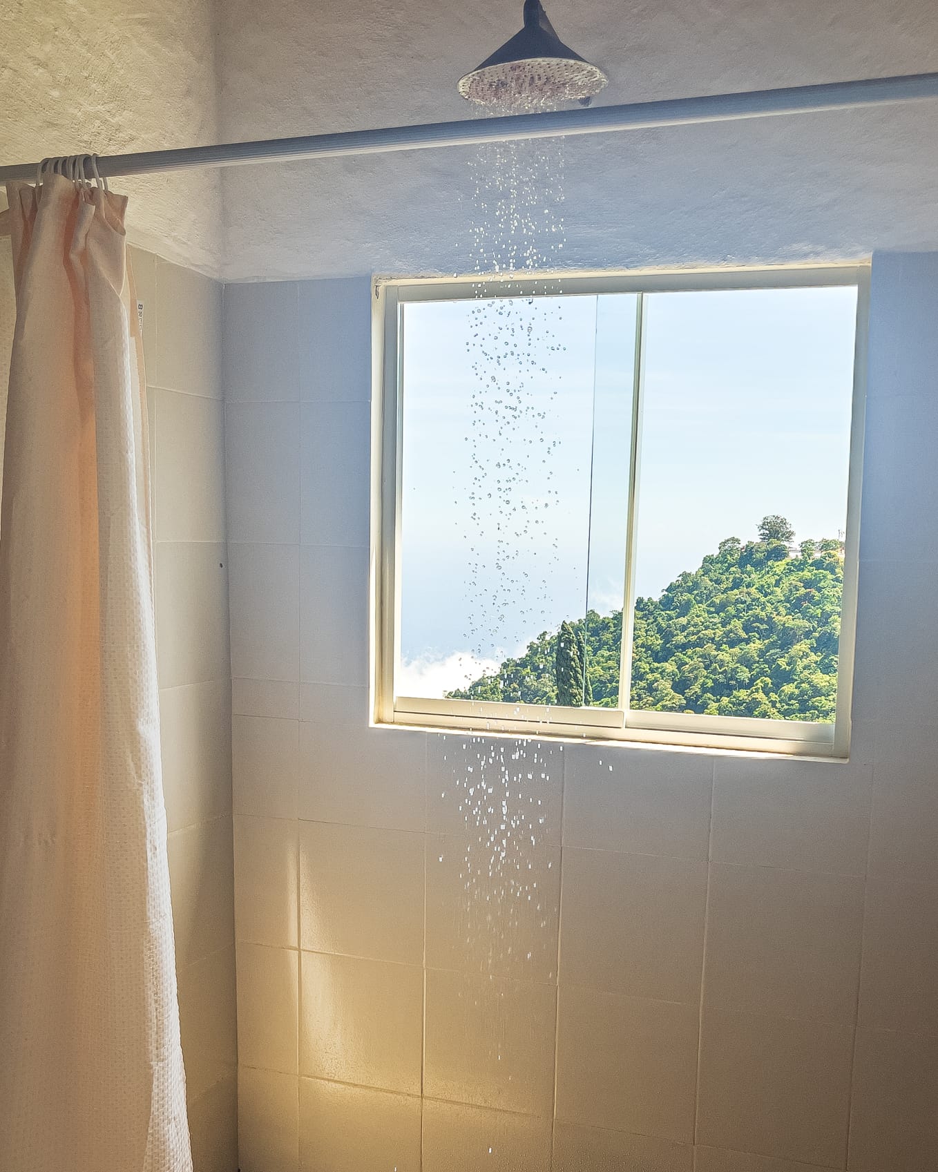 Agua cayendo de la regadera frente a la ventana con vista al monte verde y las nubes