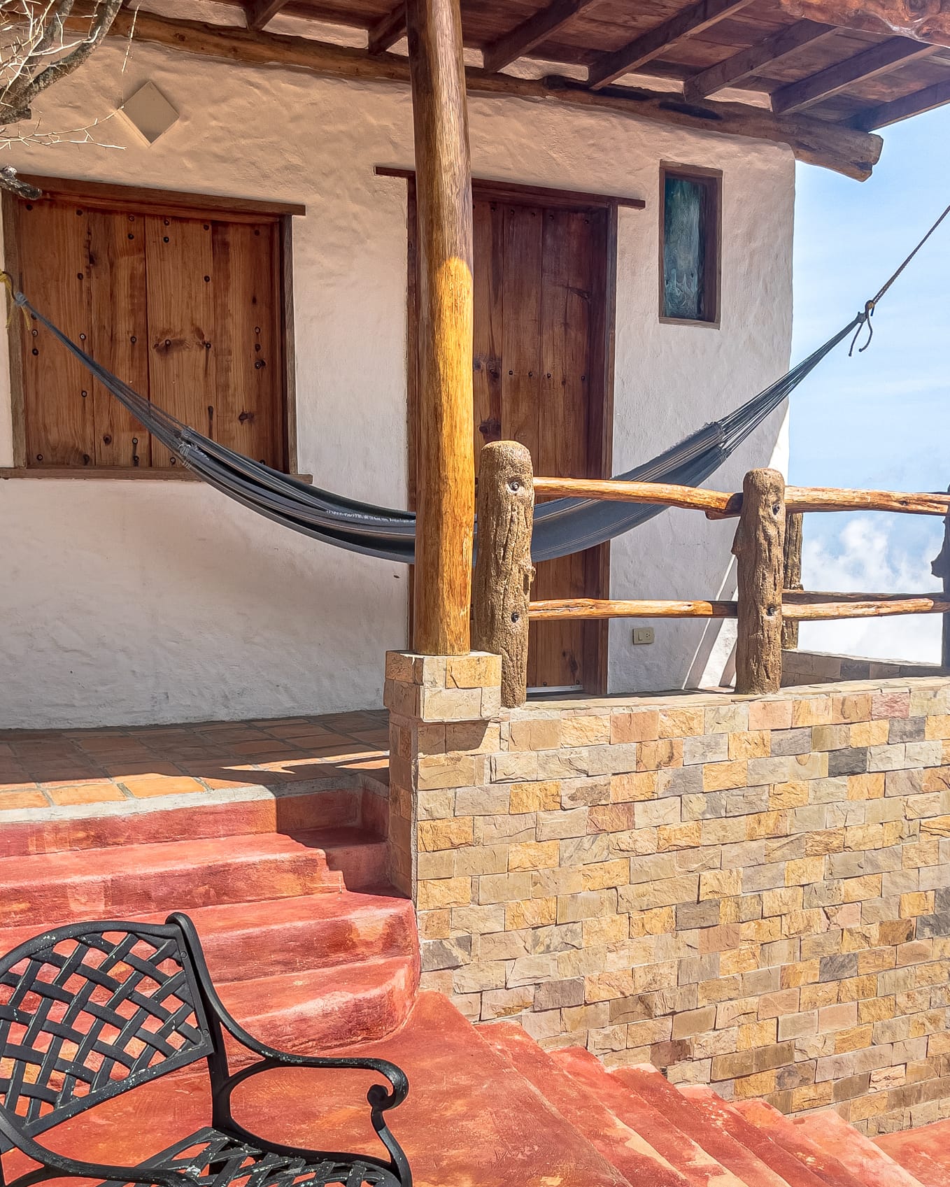 Hamaca colgada en el porche de la cabaña, entre columnas de tronco y el murito de piedra