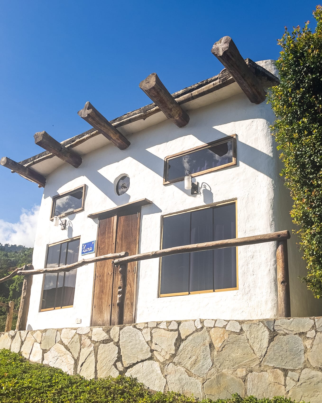 Fachada blanca de la cabaña con vigas de madera salientes, base de piedra y baranda rústica bajo un cielo despejado