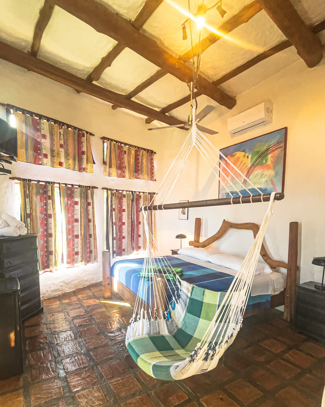 Habitación principal con cama king de troncos, silla hamaca a cuadros verdes, aire acondicionado y cortinas de colores