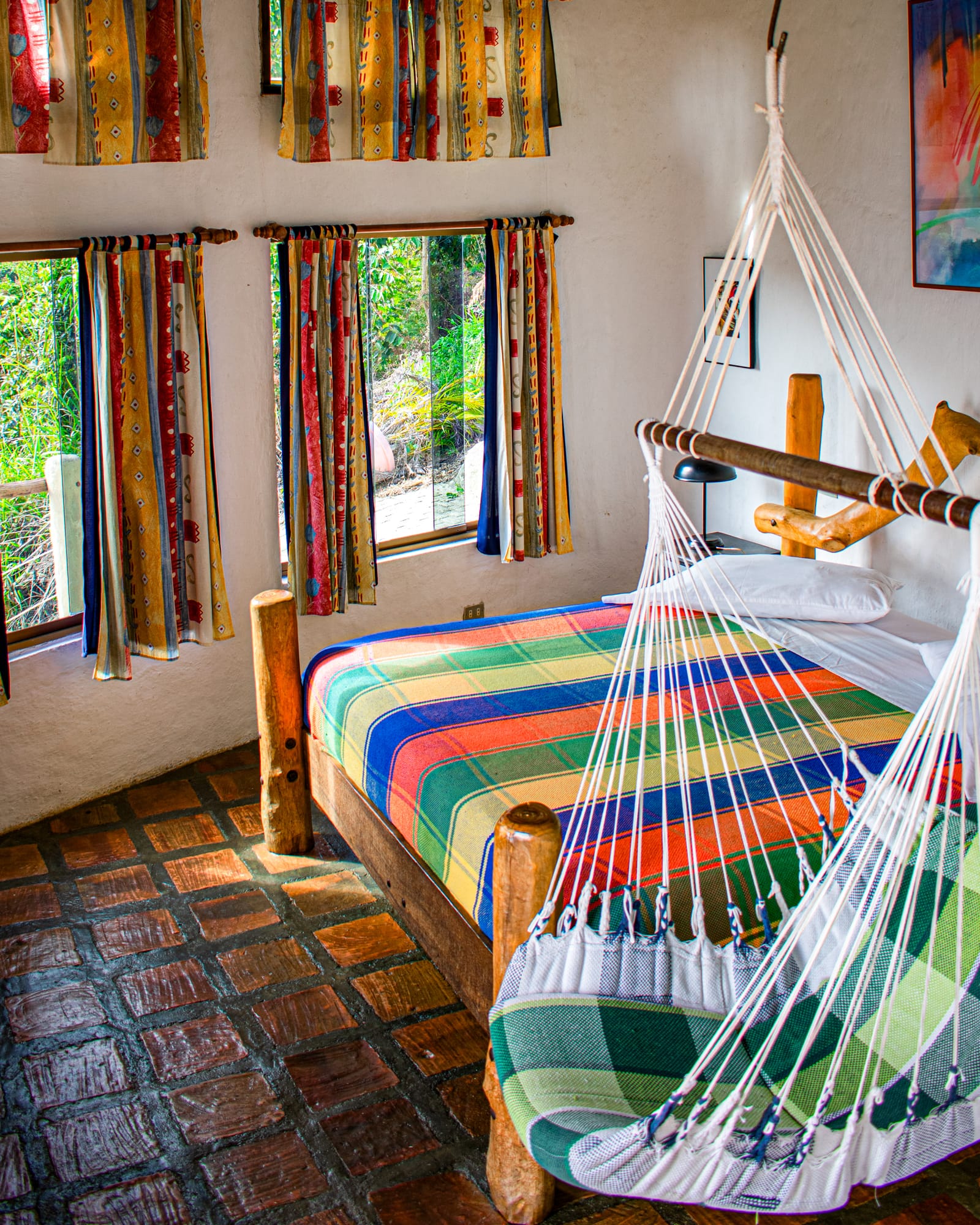 Cama king con sobrecama de cuadros vivos, hamaca a los pies y el verde de Galipán asomando por las ventanas