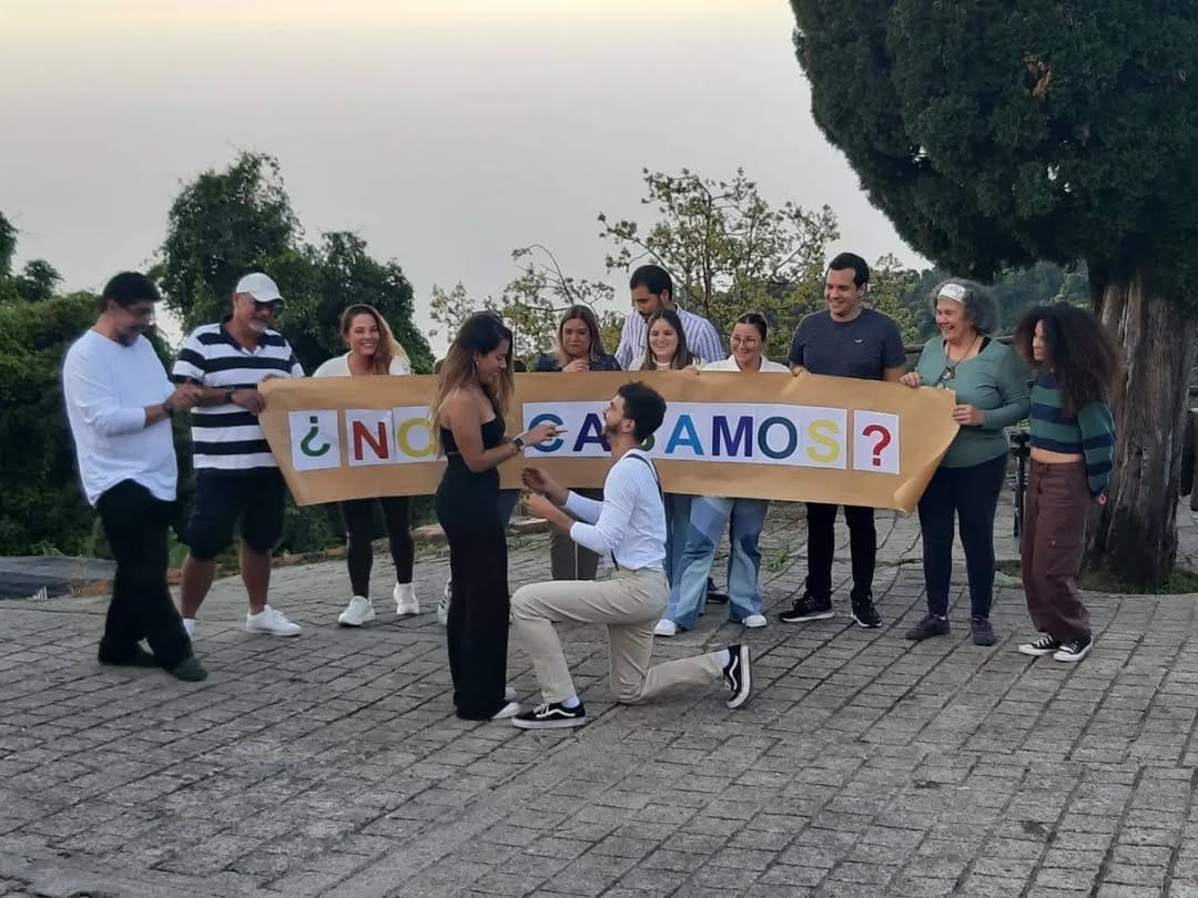 Hombre de rodillas pide matrimonio mientras sus amigos sostienen un cartel de ¿Nos casamos? al atardecer en Galipán.