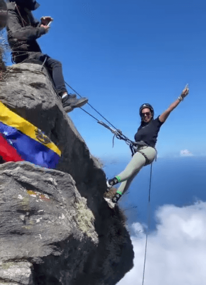 Mujer con casco y arnés descolgándose de una pared de roca con una bandera de Venezuela y nubes abajo, Picacho de Galipán