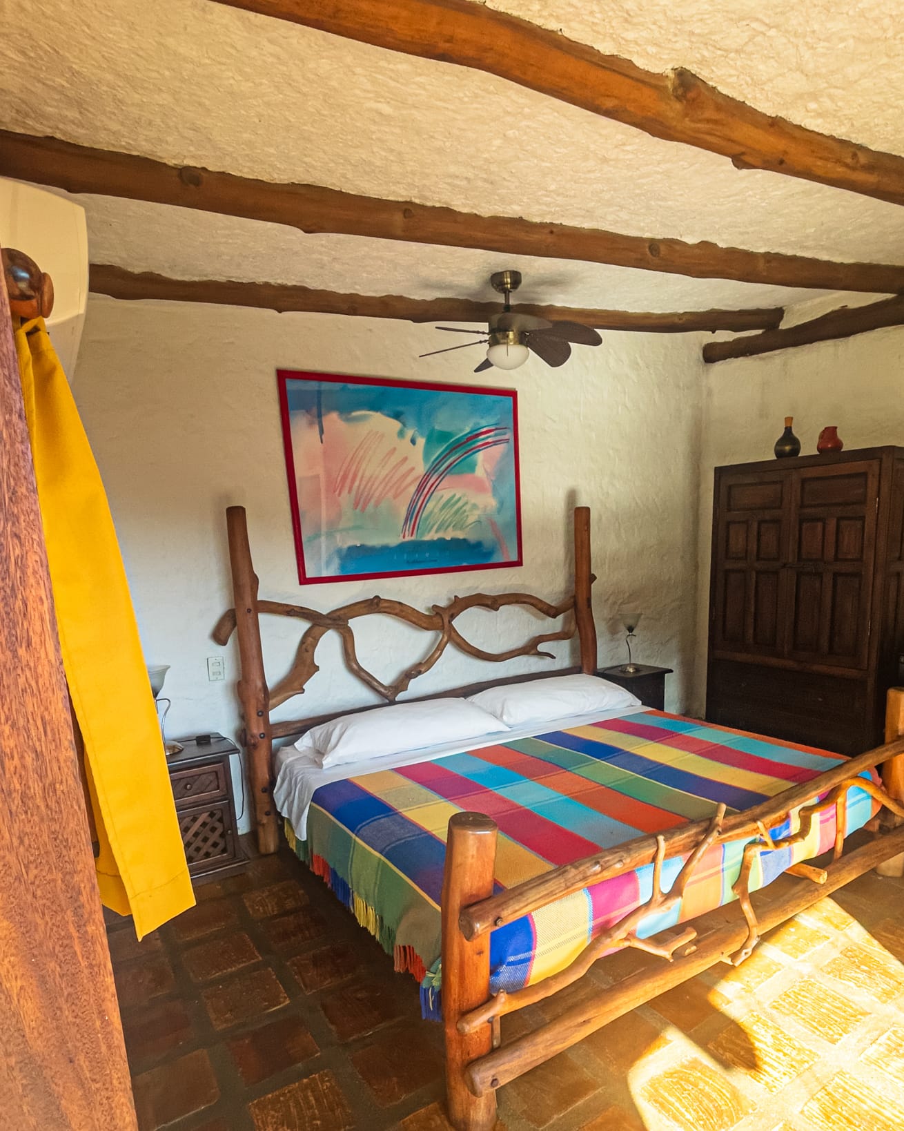 Cama matrimonial de troncos con cubrecama a rayas de colores, cuadro vivo en la pared y ventilador de techo