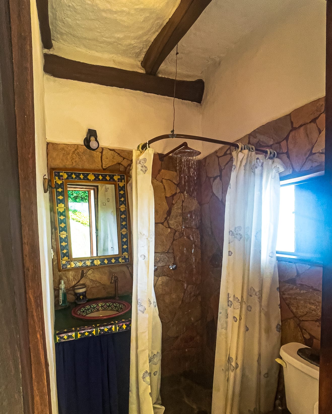 Baño con ducha de pared de piedra, cortinas blancas colgadas de un aro y lavamanos de cerámica pintada a mano