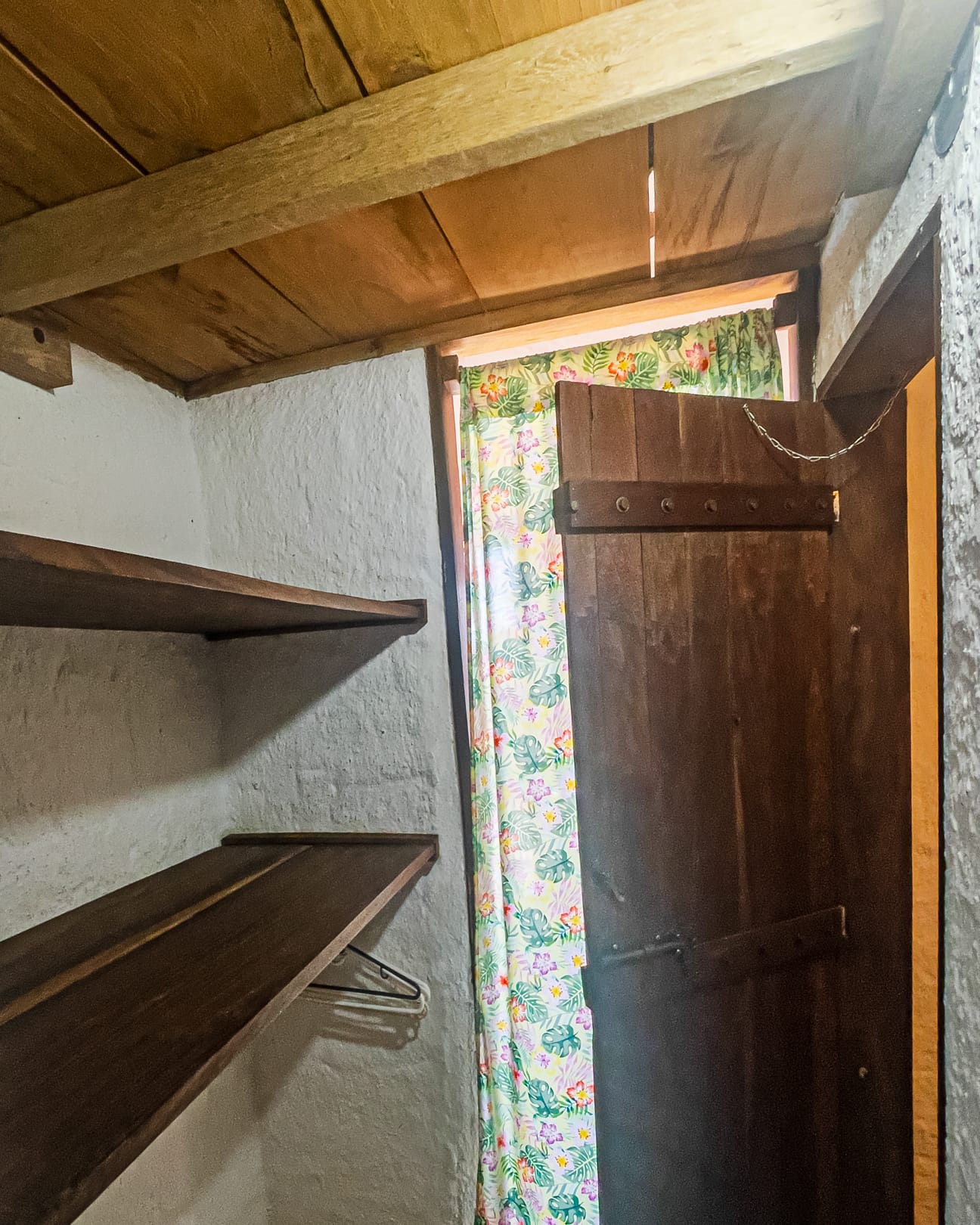 Clóset de repisas de madera oscura con tubo para colgar, junto a una cortina floreada y la puerta rústica