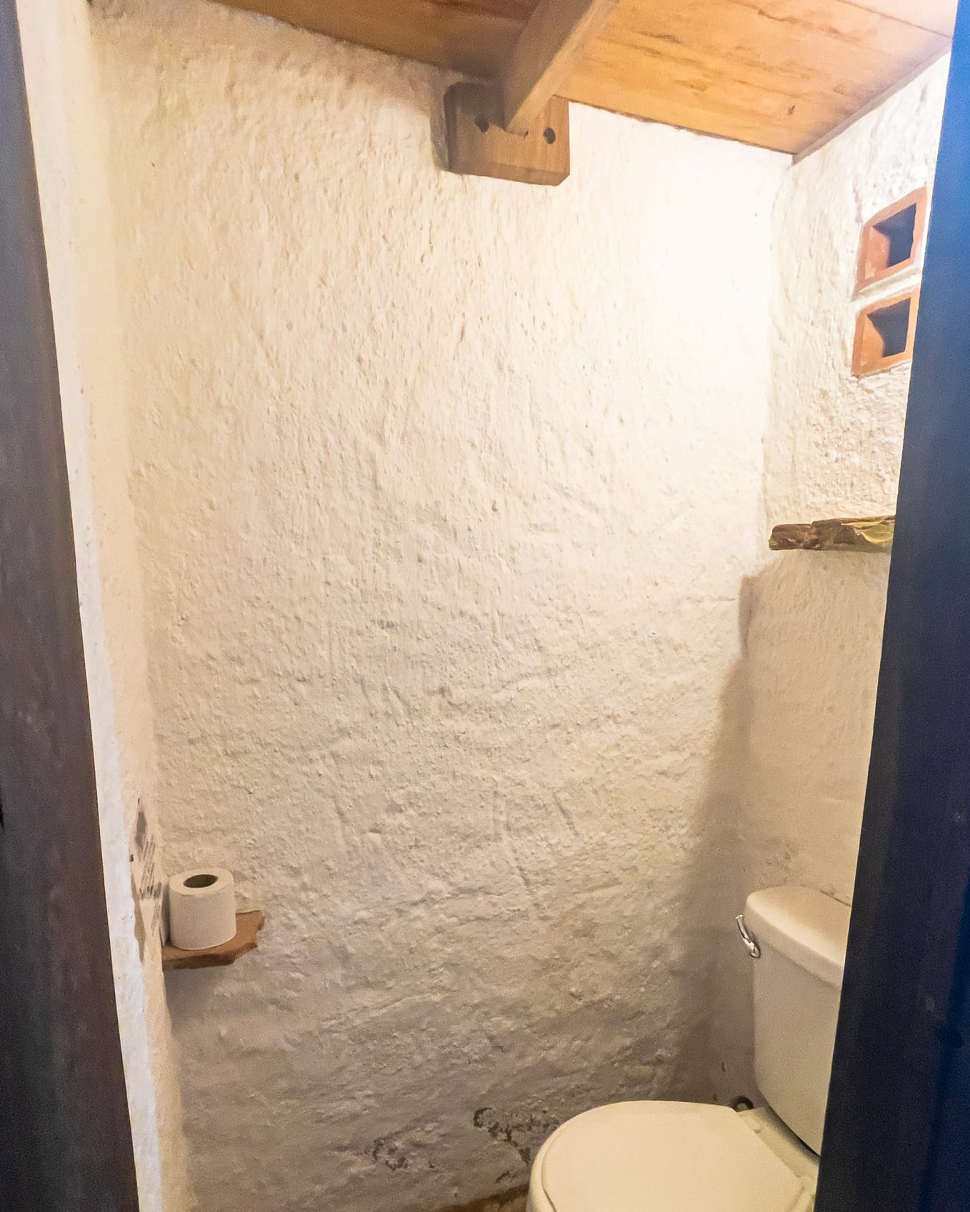 Rincón del sanitario entre paredes encaladas, con repisita de madera para el papel y bloques calados de ventilación